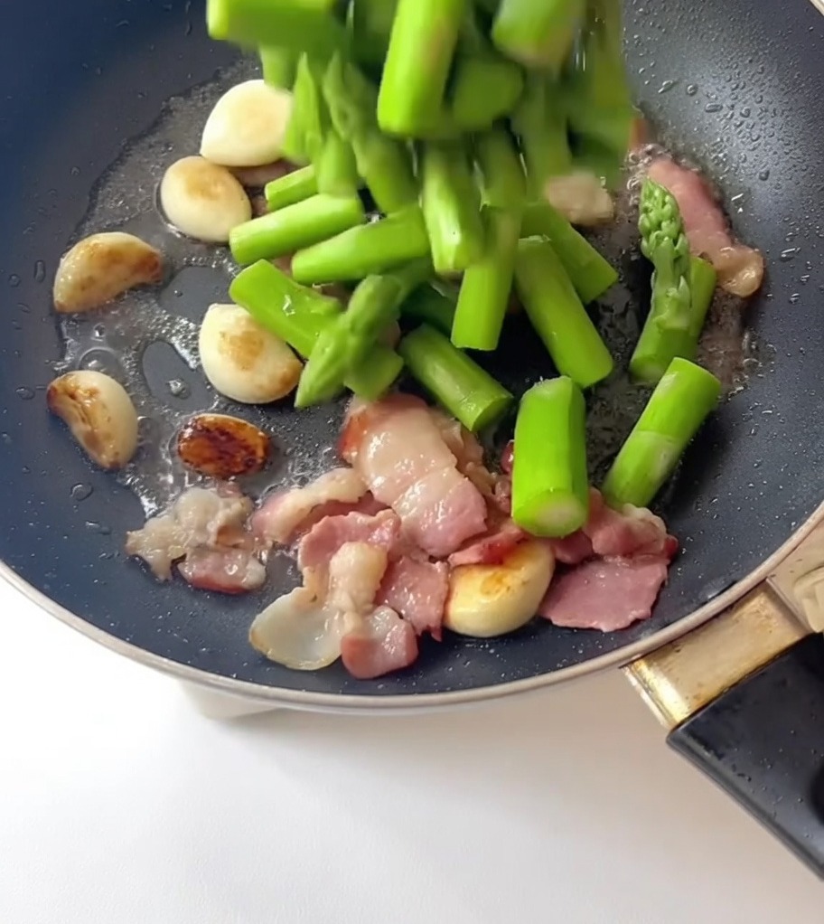 Pezzi di asparagi sbollentati versati in una padella contenente aglio e pancetta.