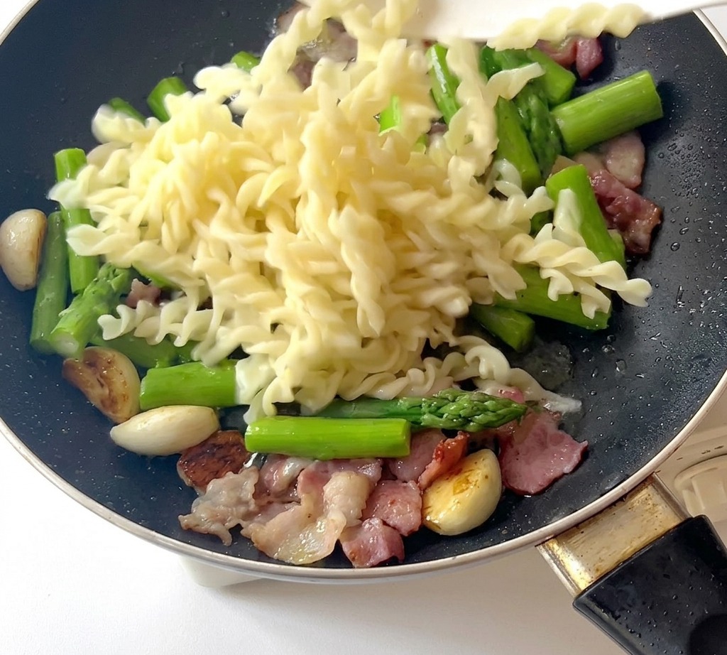 Fusilli cotti versati in una padella contenente asparagi, pancetta e aglio.