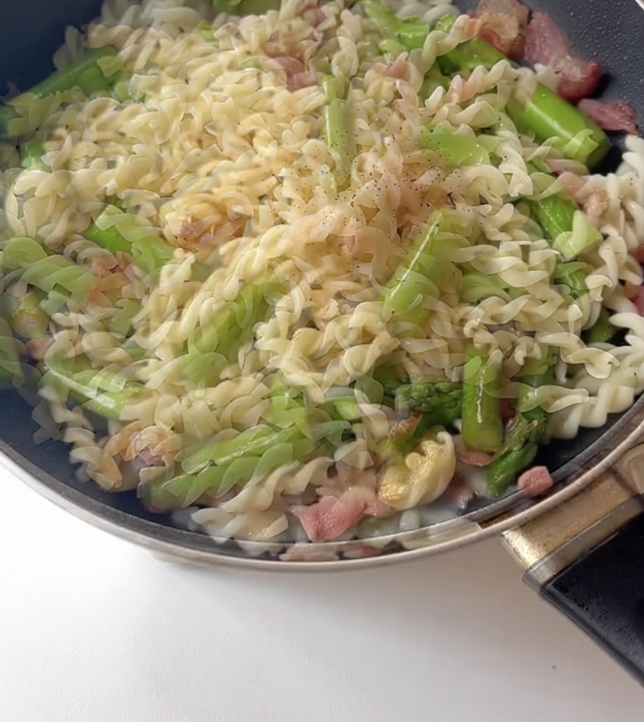 Condimenti, incluso pepe nero, spolverati su pasta, asparagi e pancetta in padella.