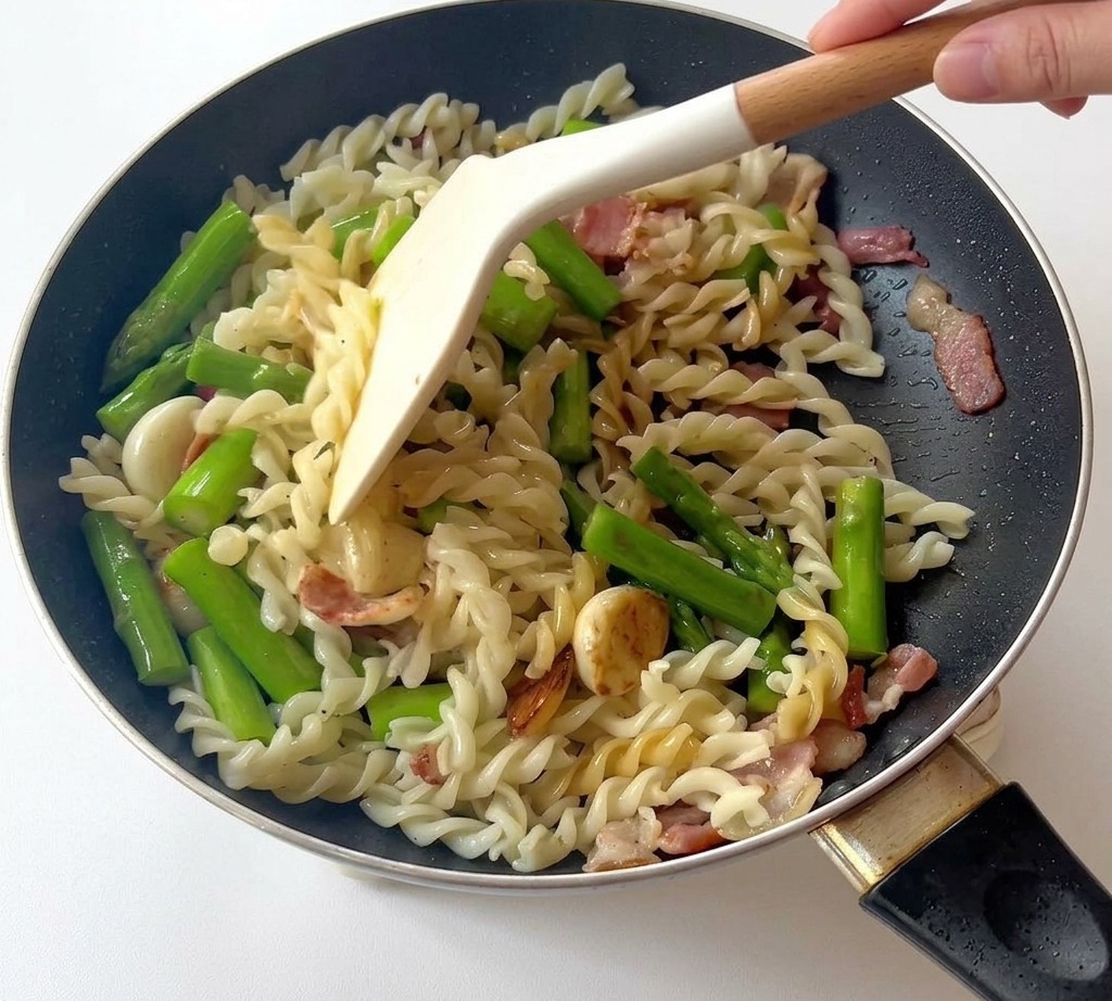 Una spatola bianca che mescola fusilli, asparagi e pancetta in una padella nera.