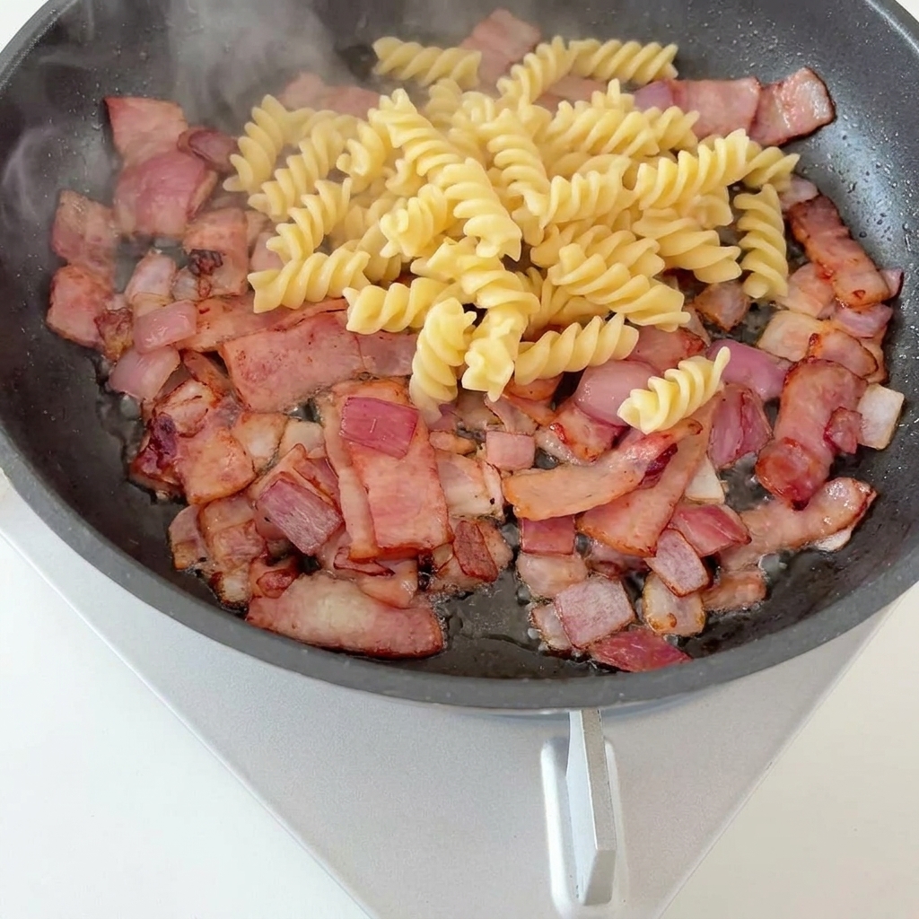 Pasta e asparagi sopra la base di pancetta e cipolle in padella.