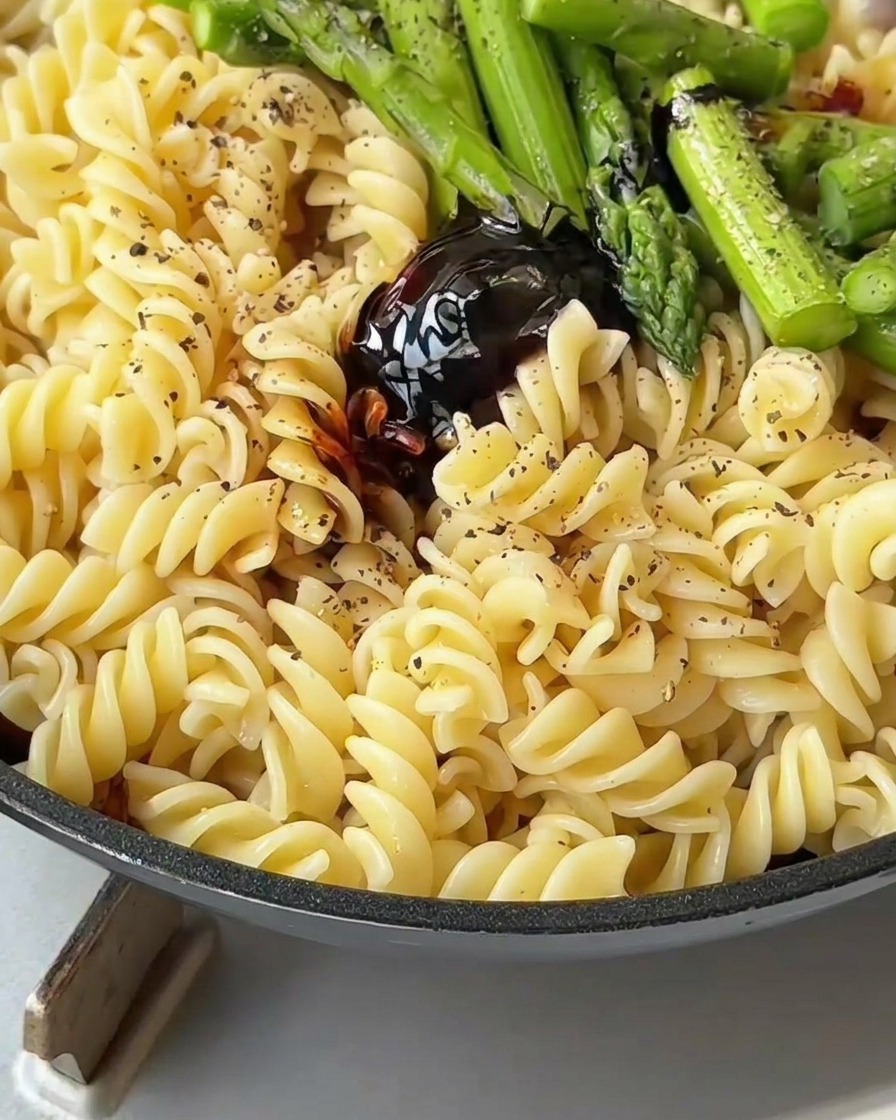 Pasta, pancetta e asparagi mescolati con una spatola in padella per ricoprirli di salsa.