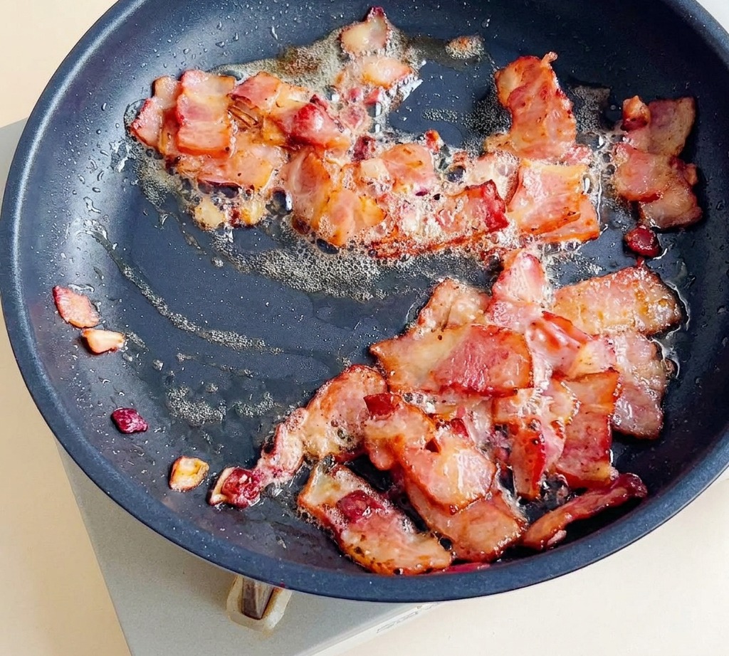 Pezzi di pancetta che friggono in padella con cipolle fino a diventare dorati e croccanti.