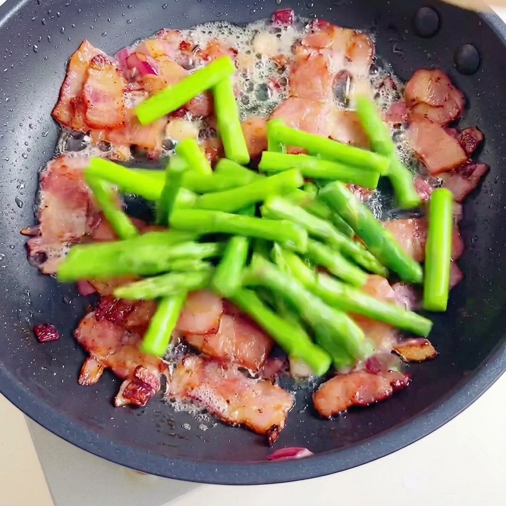 Una padella contenente pezzi di pancetta croccante e segmenti di asparagi freschi uniti ai fusilli cotti.