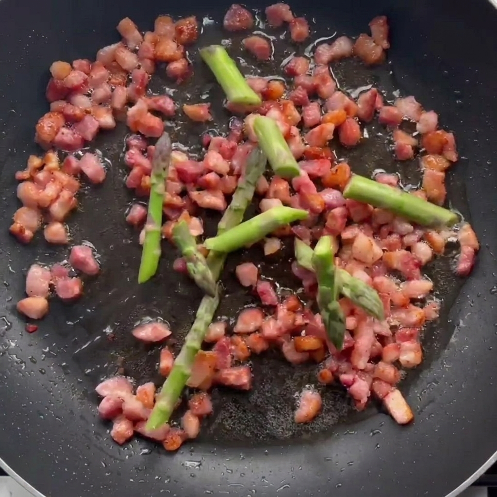 Pezzetti di asparagi che cuociono in padella insieme alla pancetta rosolata.