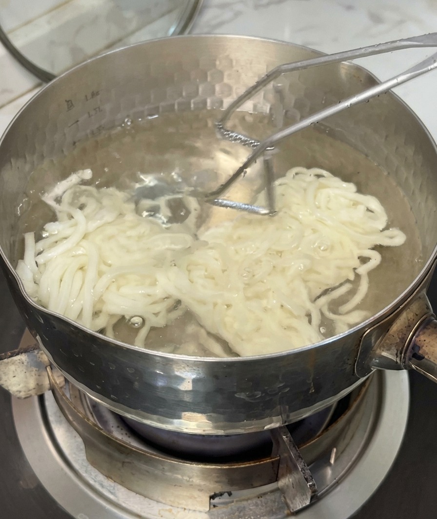 Noodles udon gommosi sollevati da una pentola dacqua bollente con uno scolapasta.