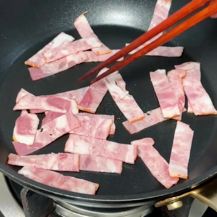 Pezzi di bacon che rosolano in padella sprigionando il grasso.