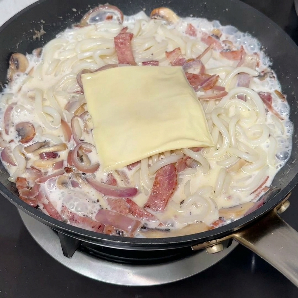 Una fetta di formaggio fuso posta sopra gli udon cremosi in padella.