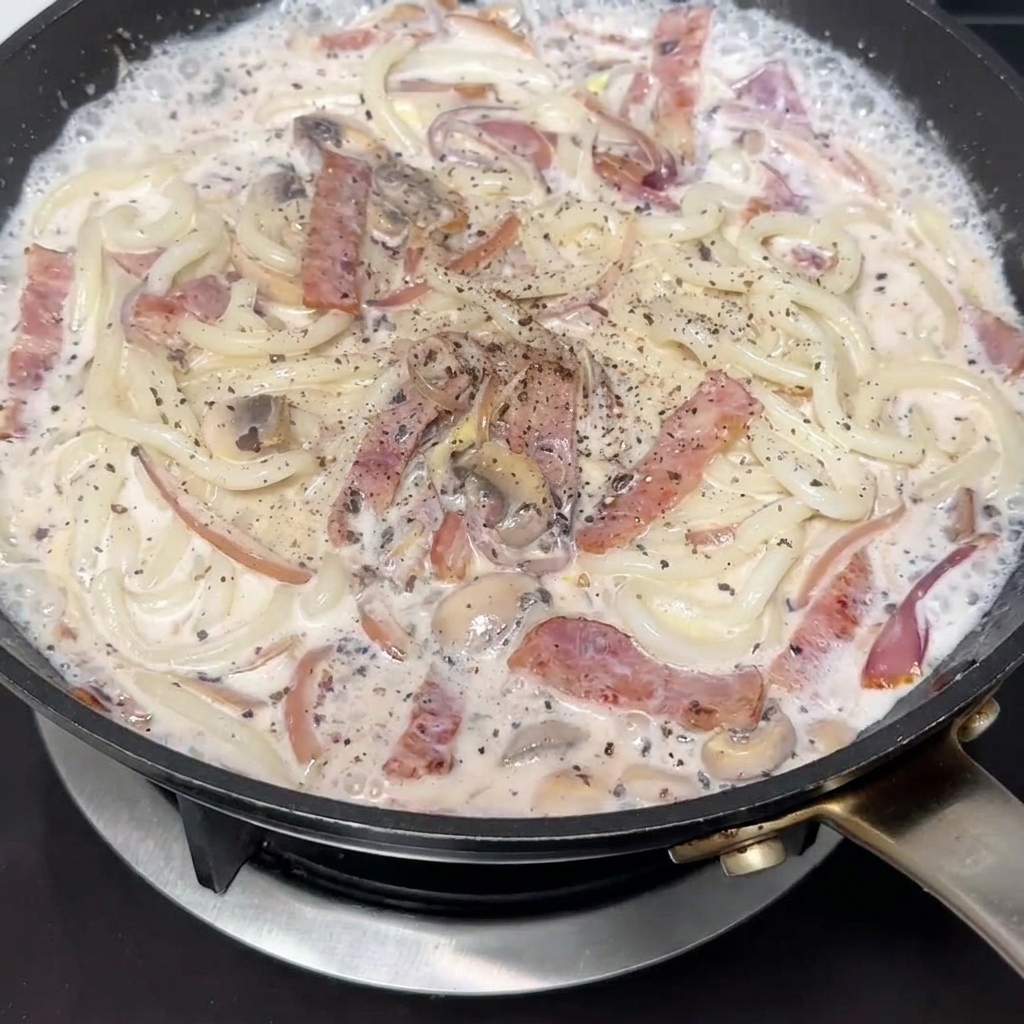 Dettaglio di pepe nero appena macinato aggiunto sugli udon cremosi.