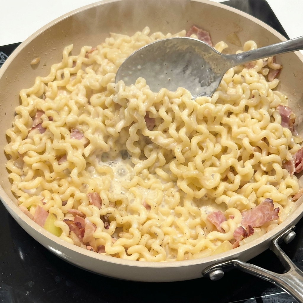 Cucchiaio di metallo che mescola pasta riccia e pancetta in una salsa cremosa in padella.