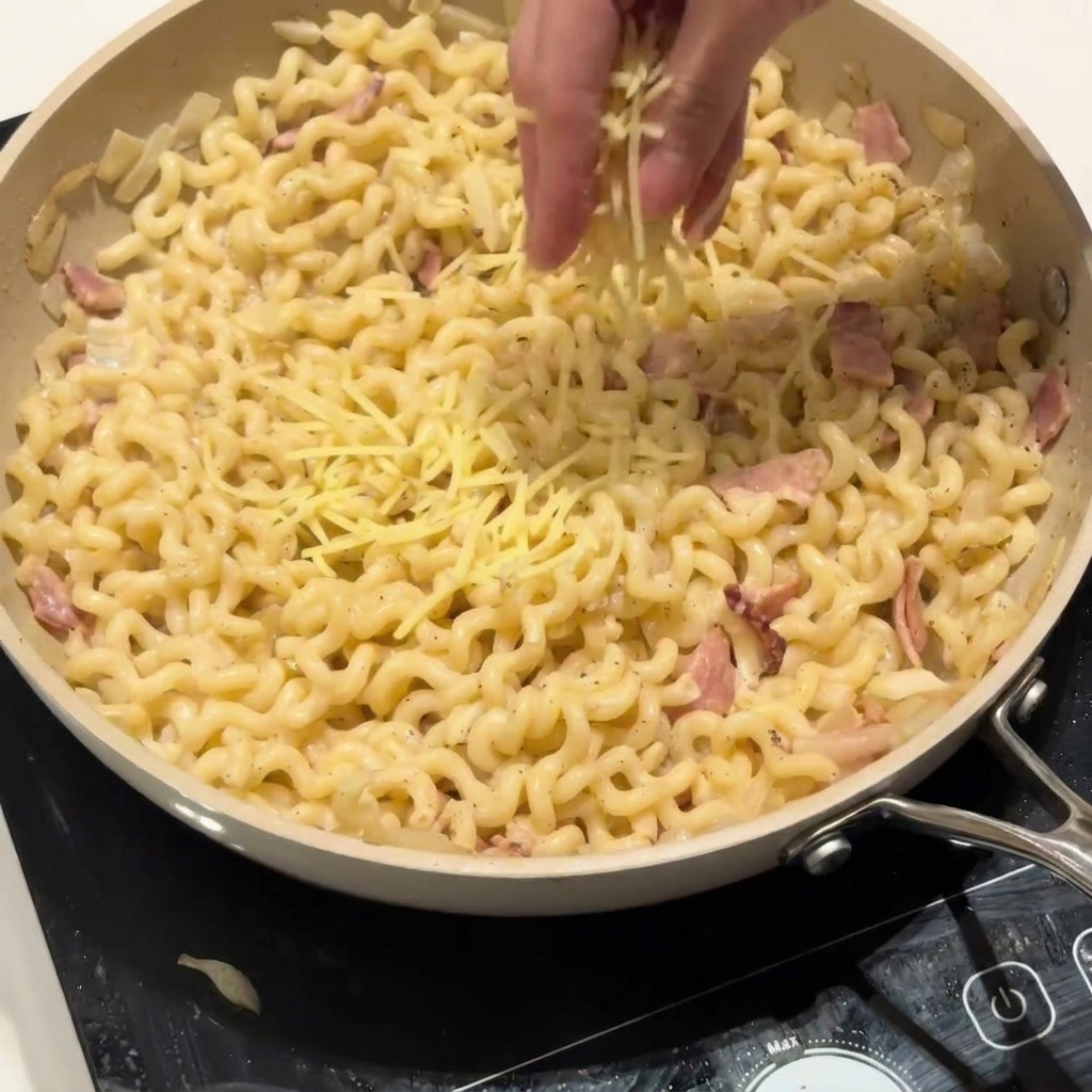 Una mano che spolverizza del parmigiano grattugiato su una padella di pasta cremosa.