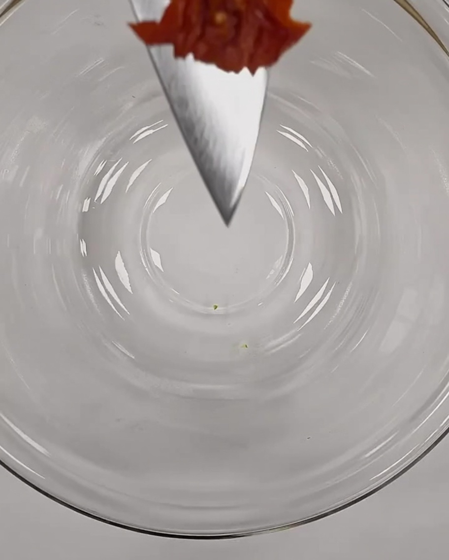 Un piccolo pezzo di pomodoro secco finemente tritato in equilibrio sulla punta di un coltello da chef sopra una ciotola di vetro.