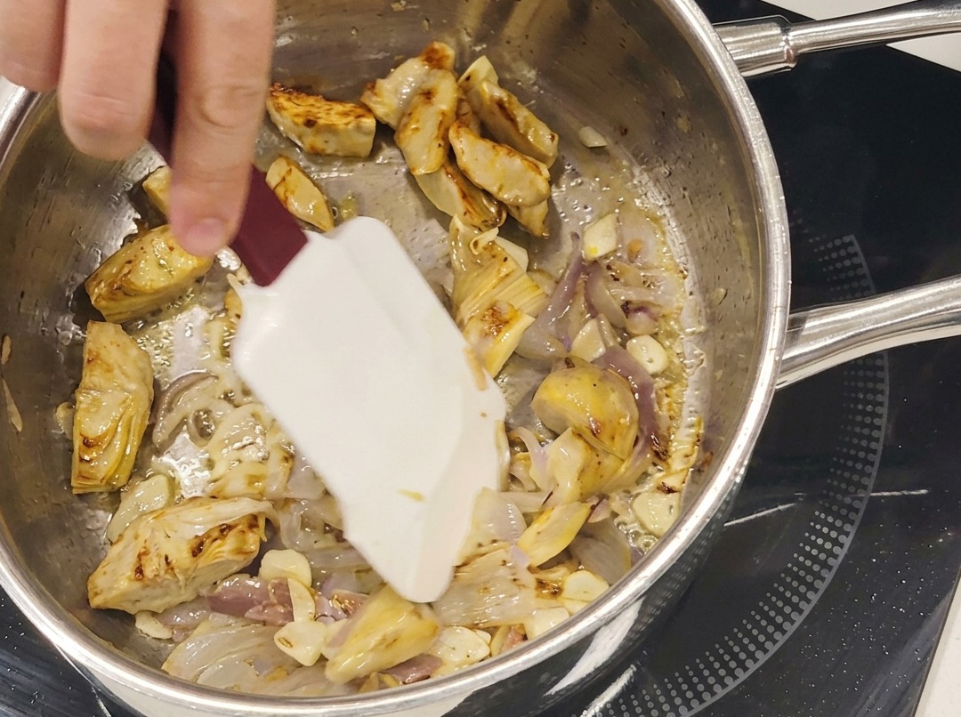 Cuori di carciofo rosolati, fettine daglio e acciughe che cuociono insieme in una padella in acciaio inox.