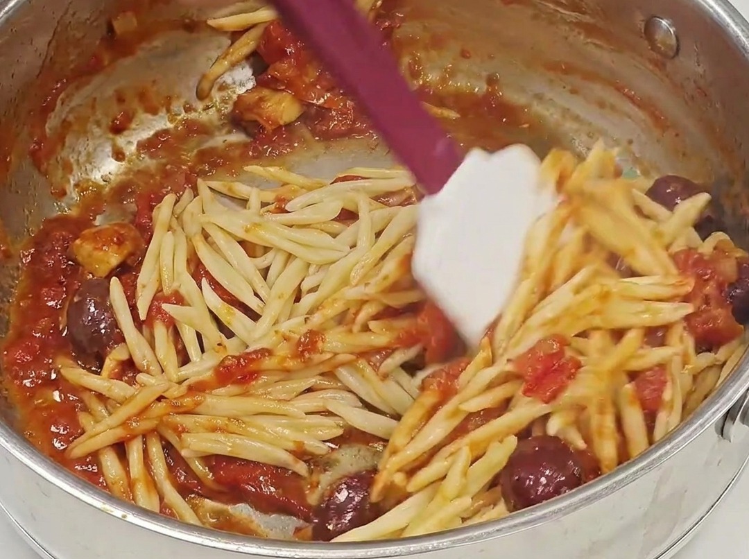 Trofie scolate aggiunte in una padella piena di ricco sugo rosso alla puttanesca.