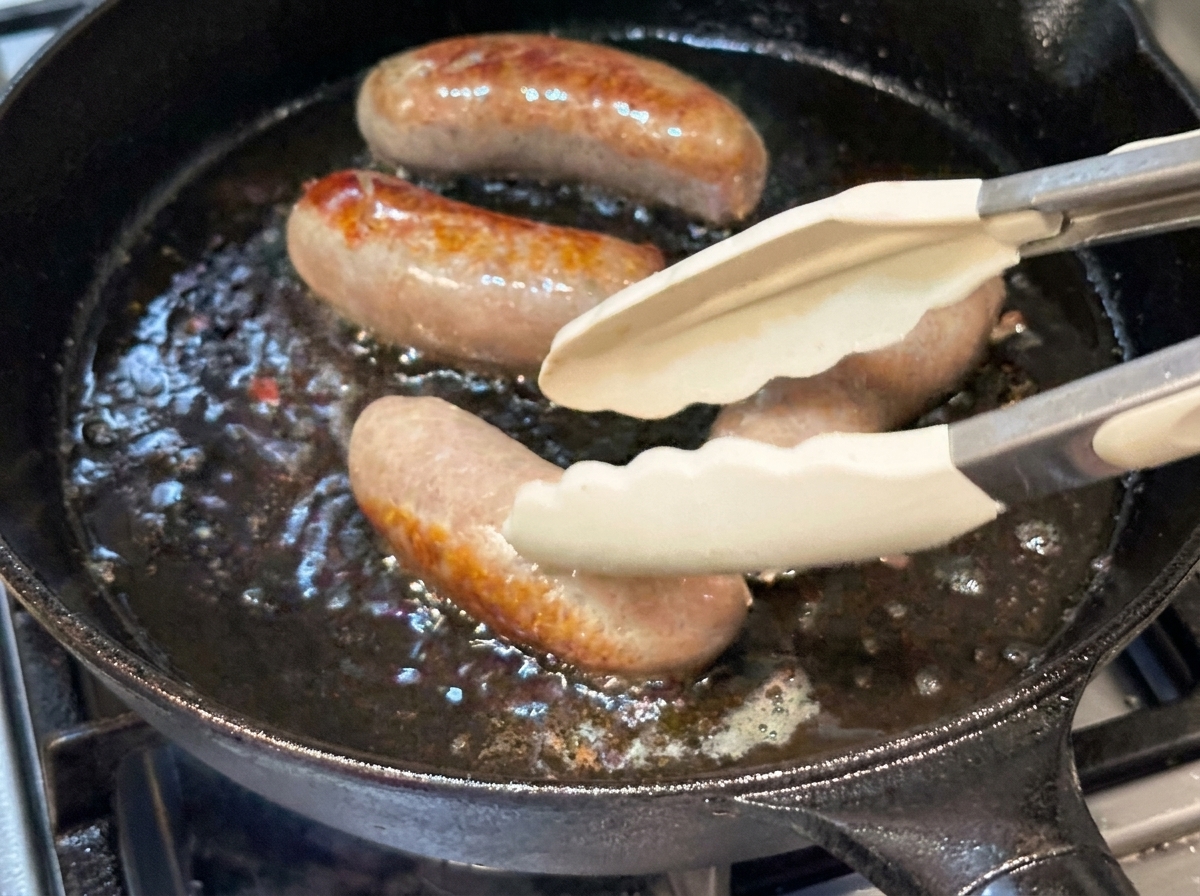 Salsicce intere girate con pinze in silicone in una padella di ghisa.