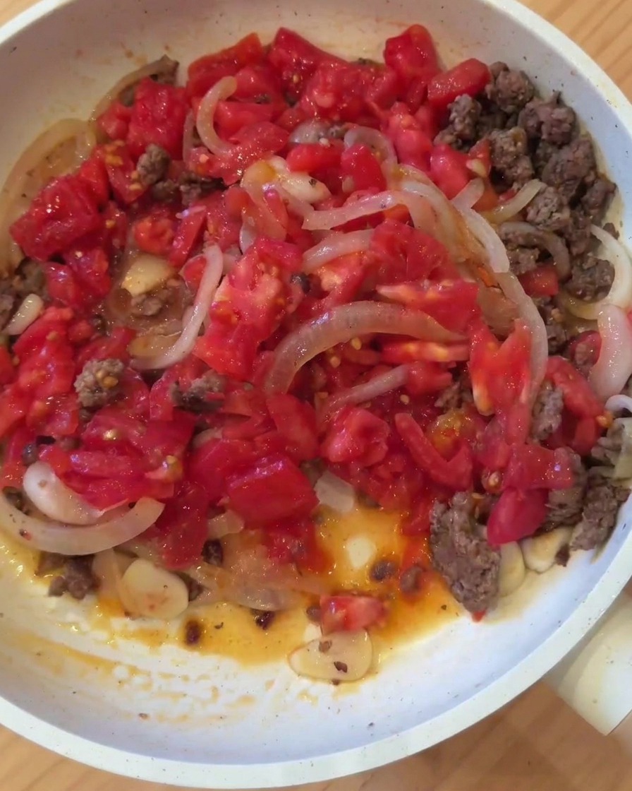 Una padella contenente carne macinata, cipolle, aglio e pomodori a pezzetti uniti sul fuoco.