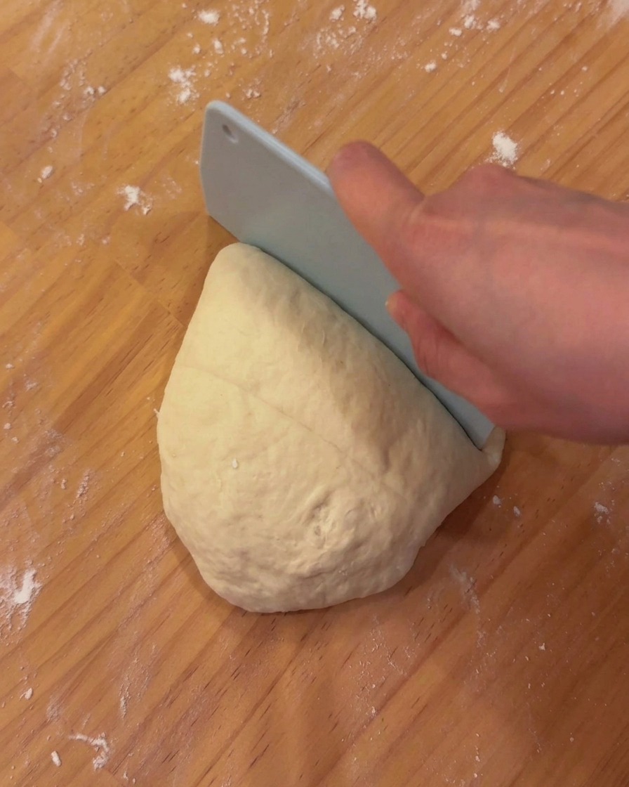 Una massa porzionata di impasto per pizza liscio e fermentato che riposa su un asse di legno con un raschietto per pasta.