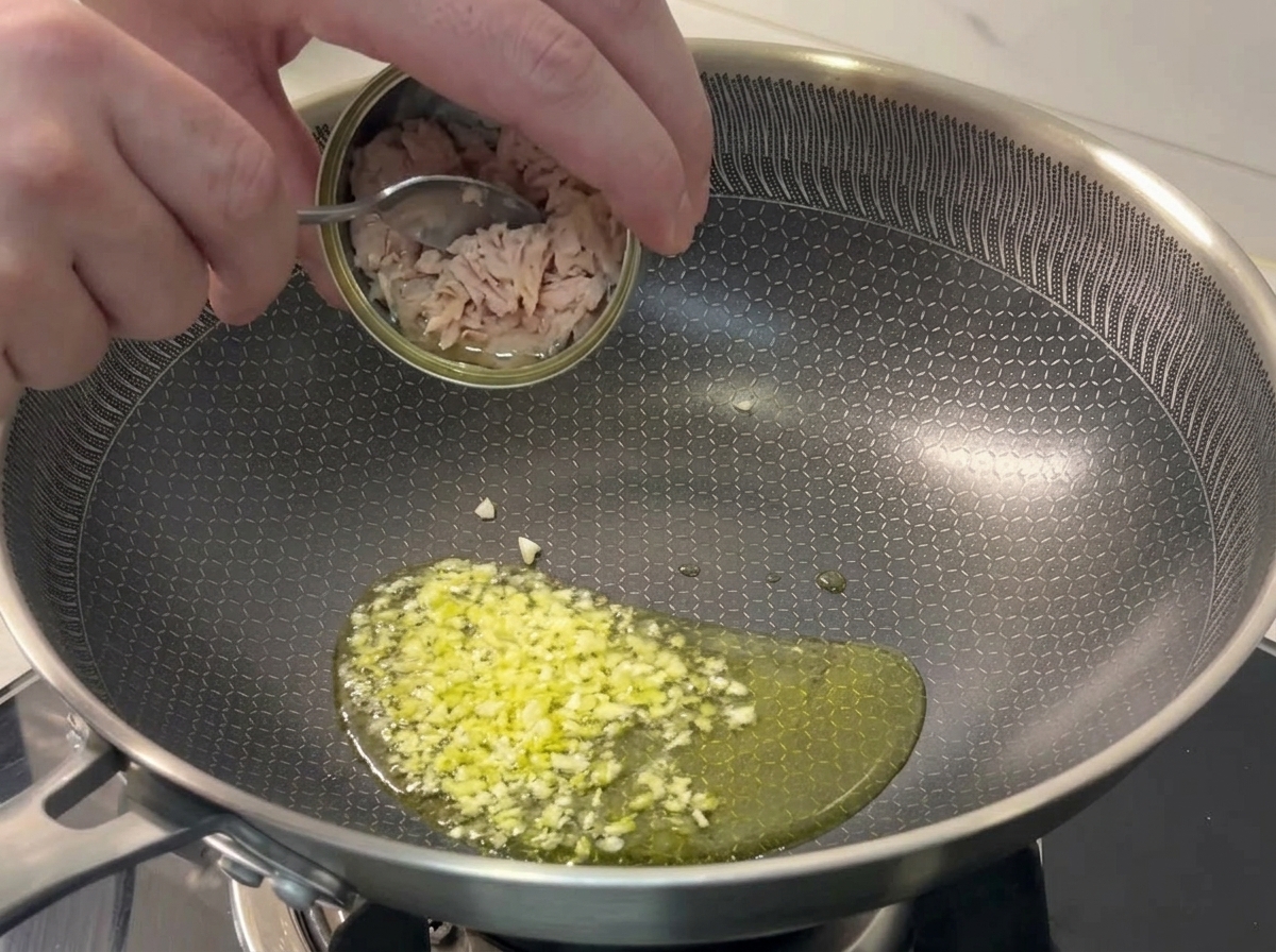 Cucchiaio che versa pezzi di tonno da una scatoletta in una padella con aglio sfrigolante.