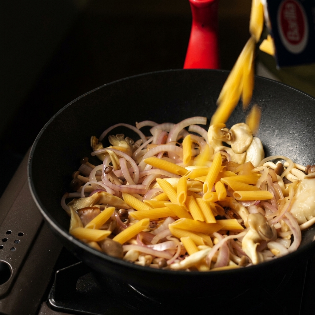 Penne secche versate da una scatola in una padella nera con funghi e cipolle saltate.