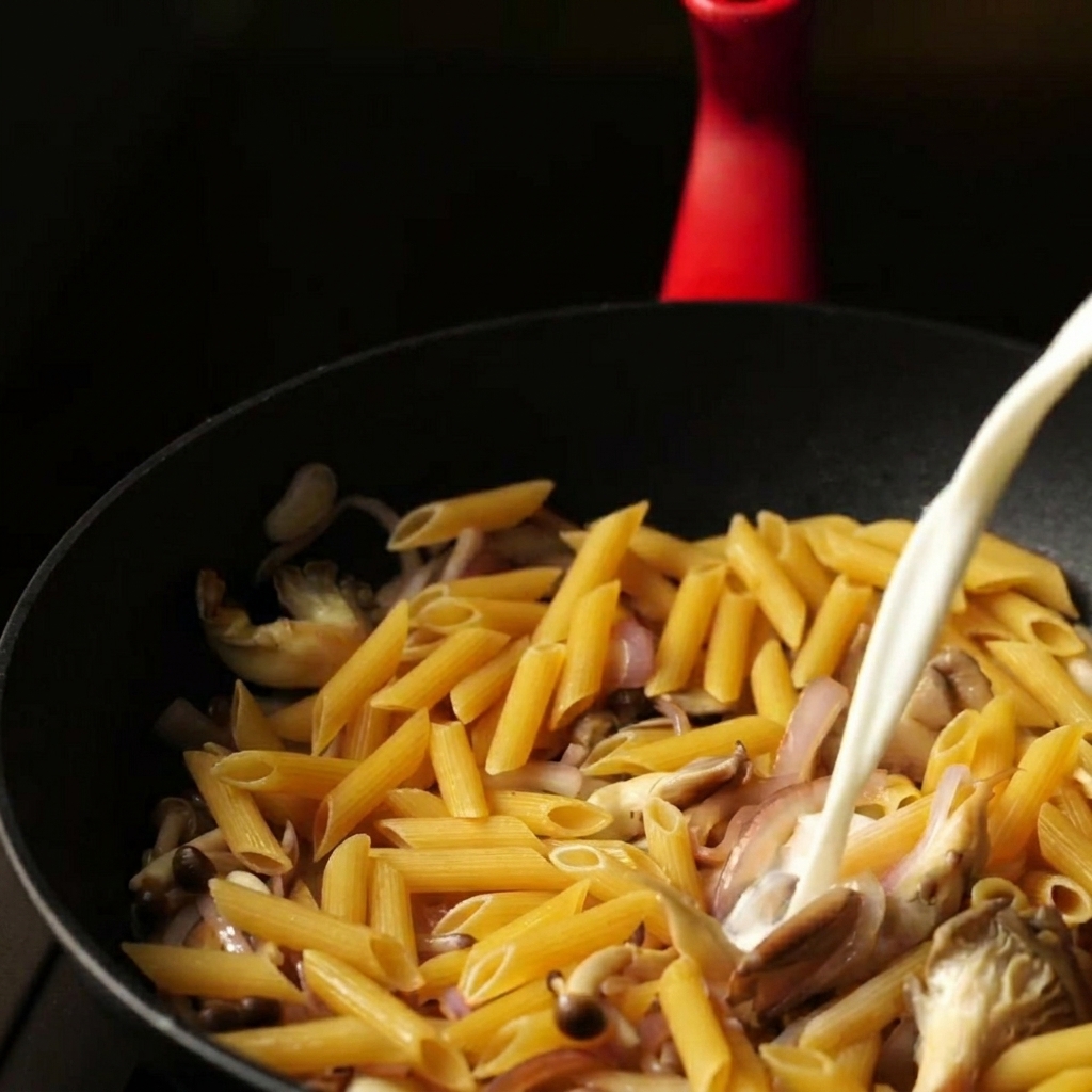 Un flusso costante di miscela di latte e acqua versato in una padella sopra penne secche e verdure saltate.