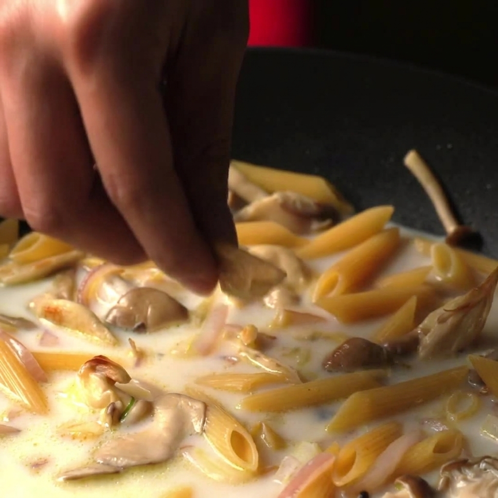 Dita che lasciano cadere un dado da brodo in una padella piena di penne che sobbollono in un brodo bianco cremoso.