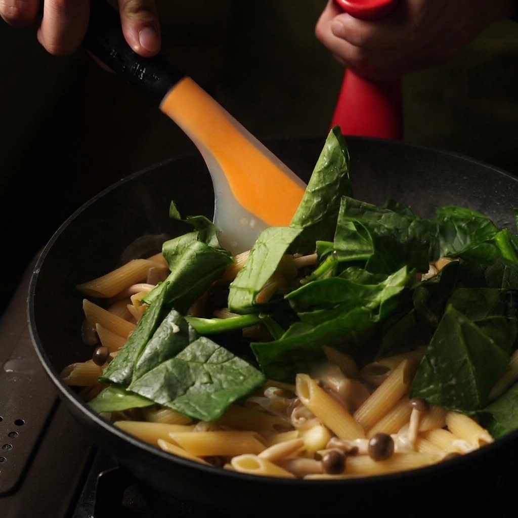 Una spatola arancione che mescola foglie di spinaci freschi verdi e burro in una padella di penne cremose cotte.