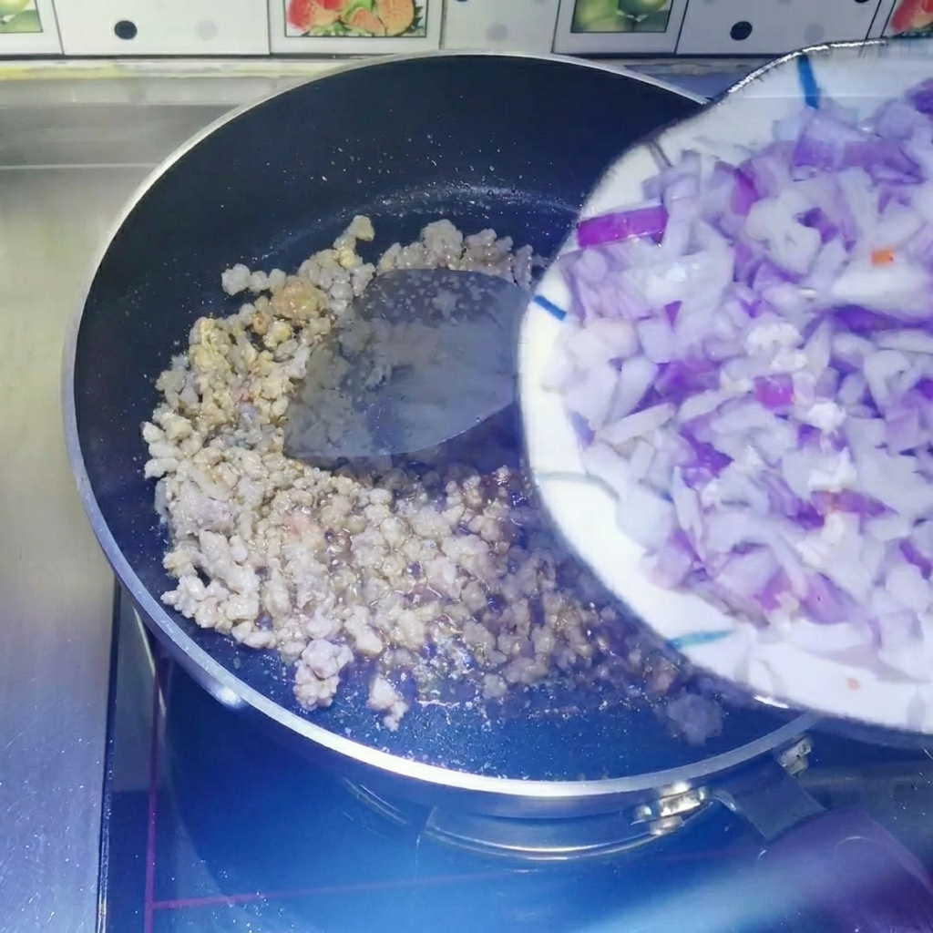 Cipolle rosse a cubetti fatte scivolare da un piatto bianco in una padella antiaderente contenente la carne macinata cotta.