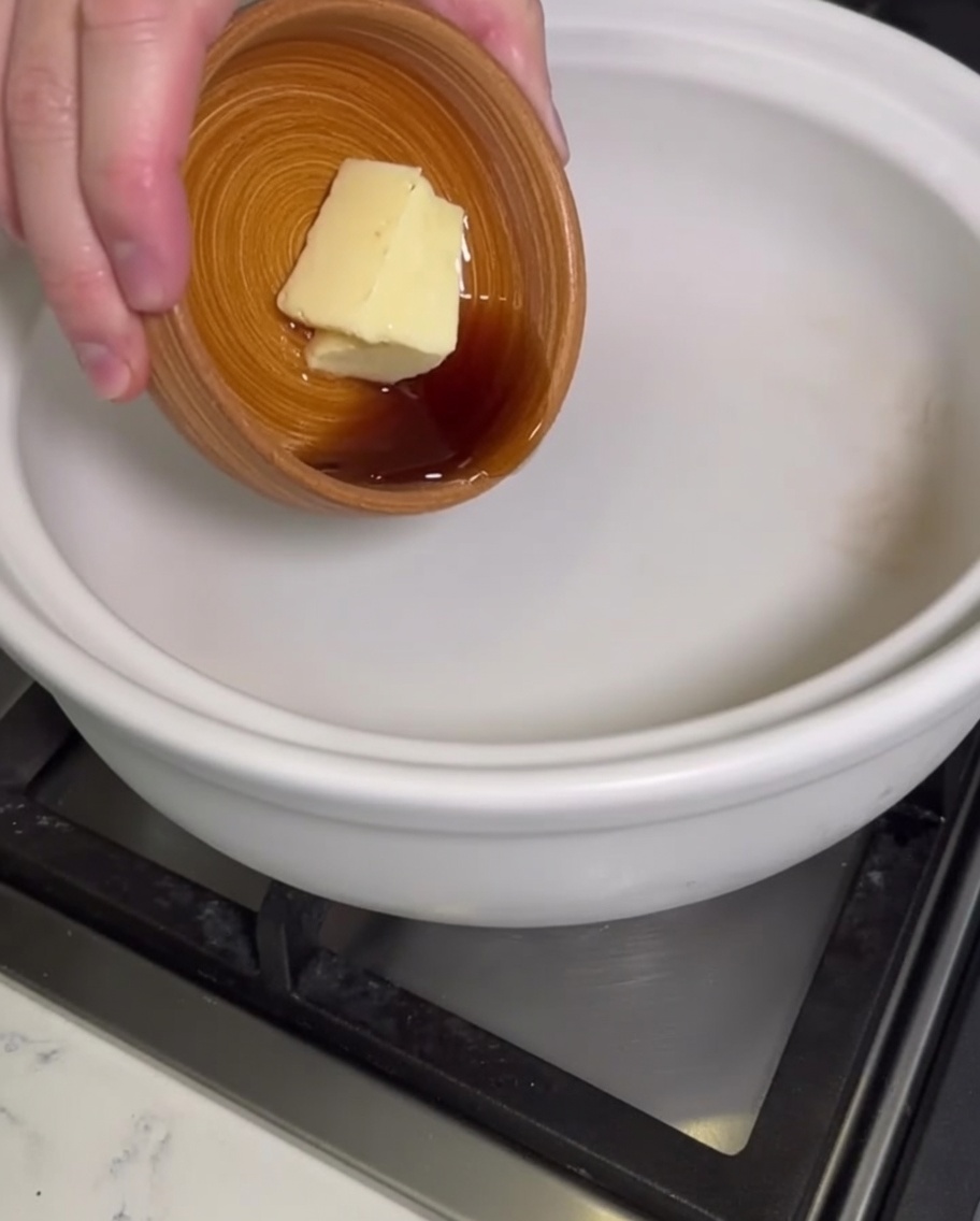 Una mano aggiunge un blocco di burro e olio di sesamo liquido da una ciotolina di legno in una pentola di terracotta bianca vuota.