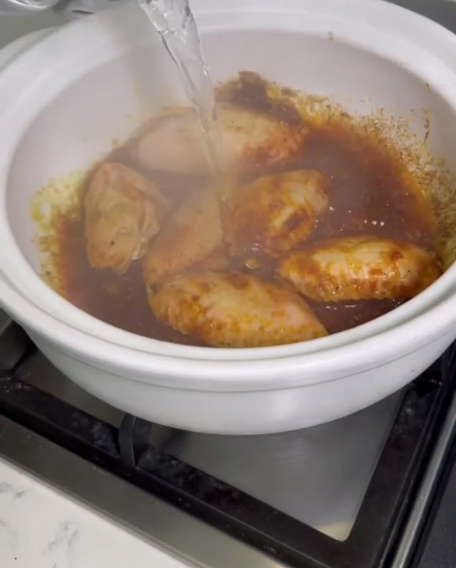 Acqua limpida versata da un recipiente in una pentola di terracotta scoppiettante piena di ali di pollo al curry.
