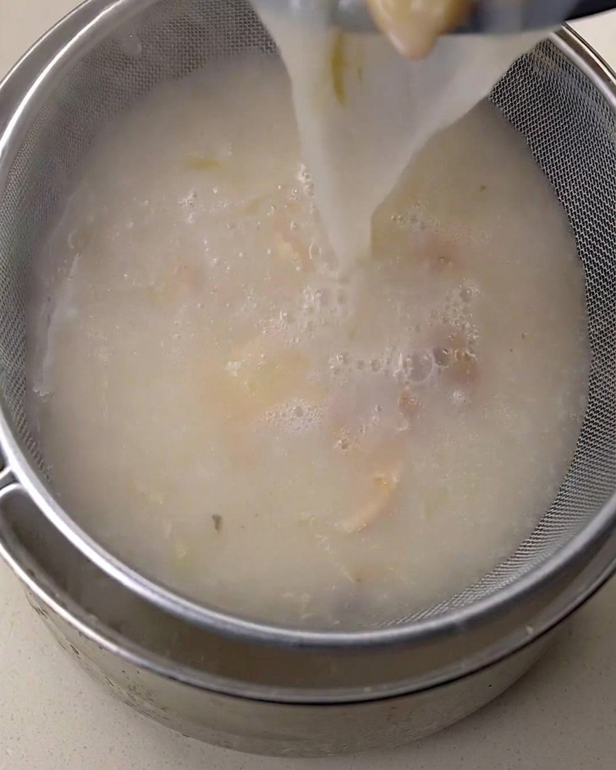 Brodo di pollo bianco e cremoso versato attraverso un colino a maglia fine in un contenitore di metallo