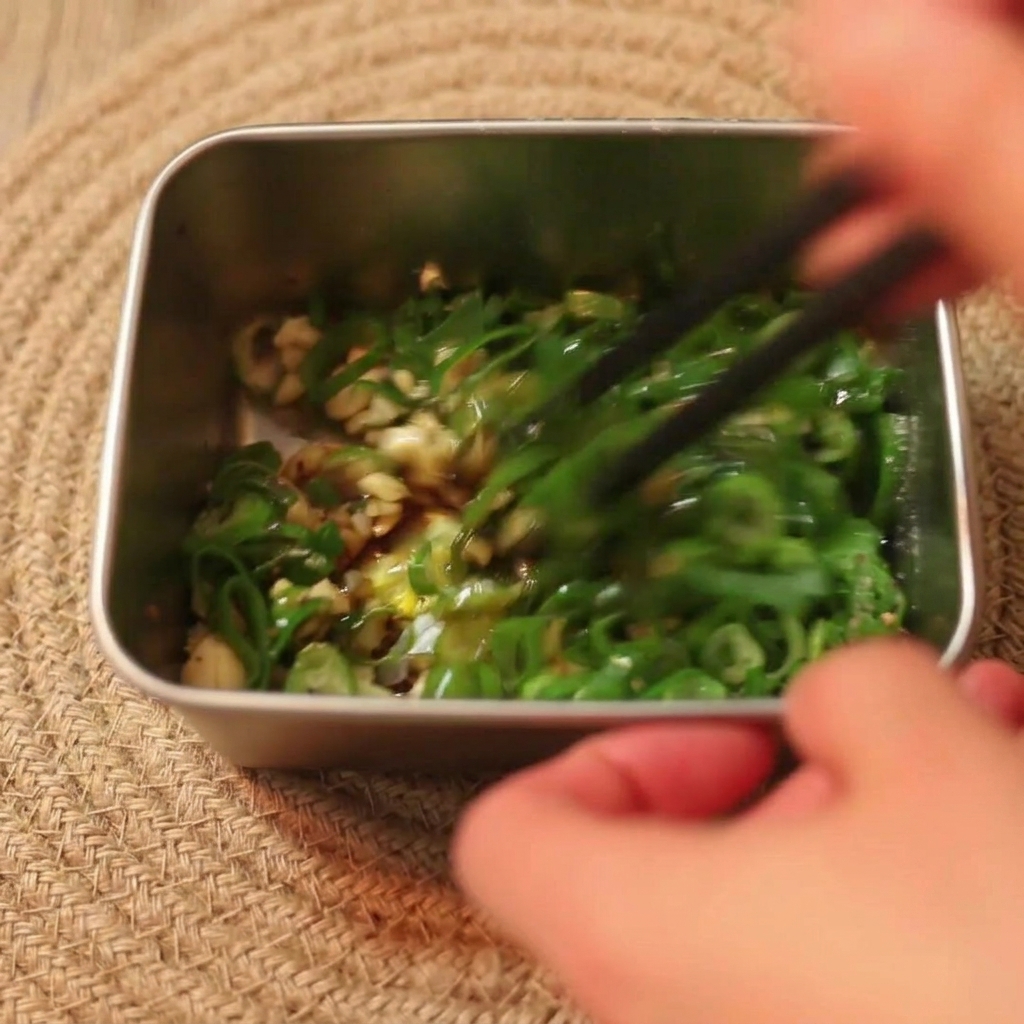 Bacchette nere che mescolano un composto di scalogno verde, aglio e salsa di soia in un contenitore di metallo.