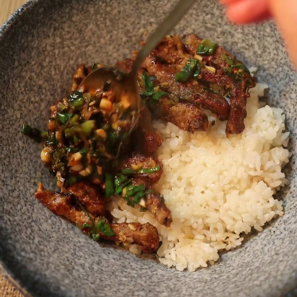 Un cucchiaio che serve manzo cotto caldo e salsa allo scalogno verde sopra un letto di riso bianco in una ciotola di ceramica.