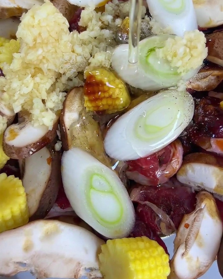 Olio versato sopra una teglia contenente manzo affettato, funghi shiitake, baby corn, cipollotti e aglio.