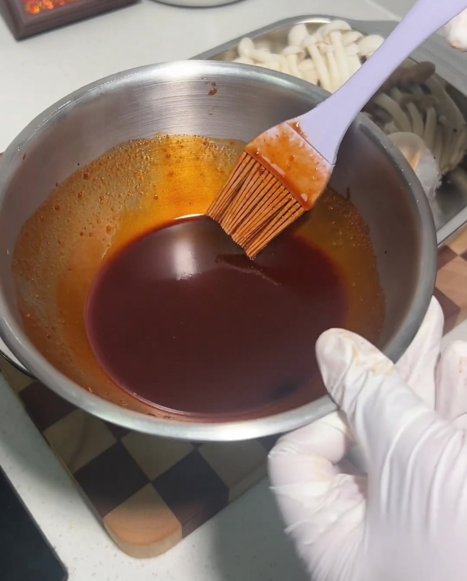 Una ciotola di metallo contenente una miscela di salsa scura e piccante che viene mescolata con un pennello in silicone.