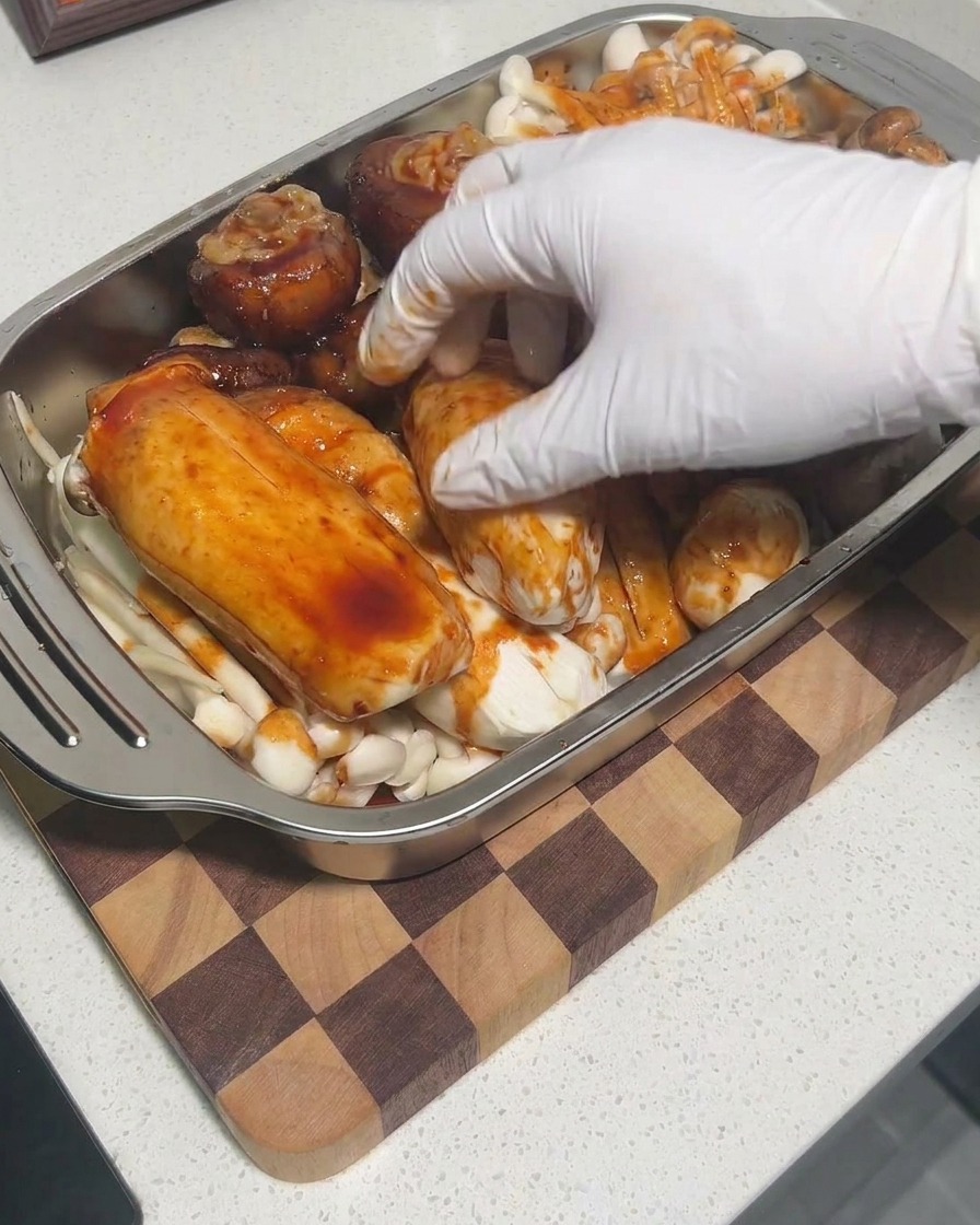 Mani con guanti che massaggiano la salsa sui funghi in una teglia per garantire una copertura completa.