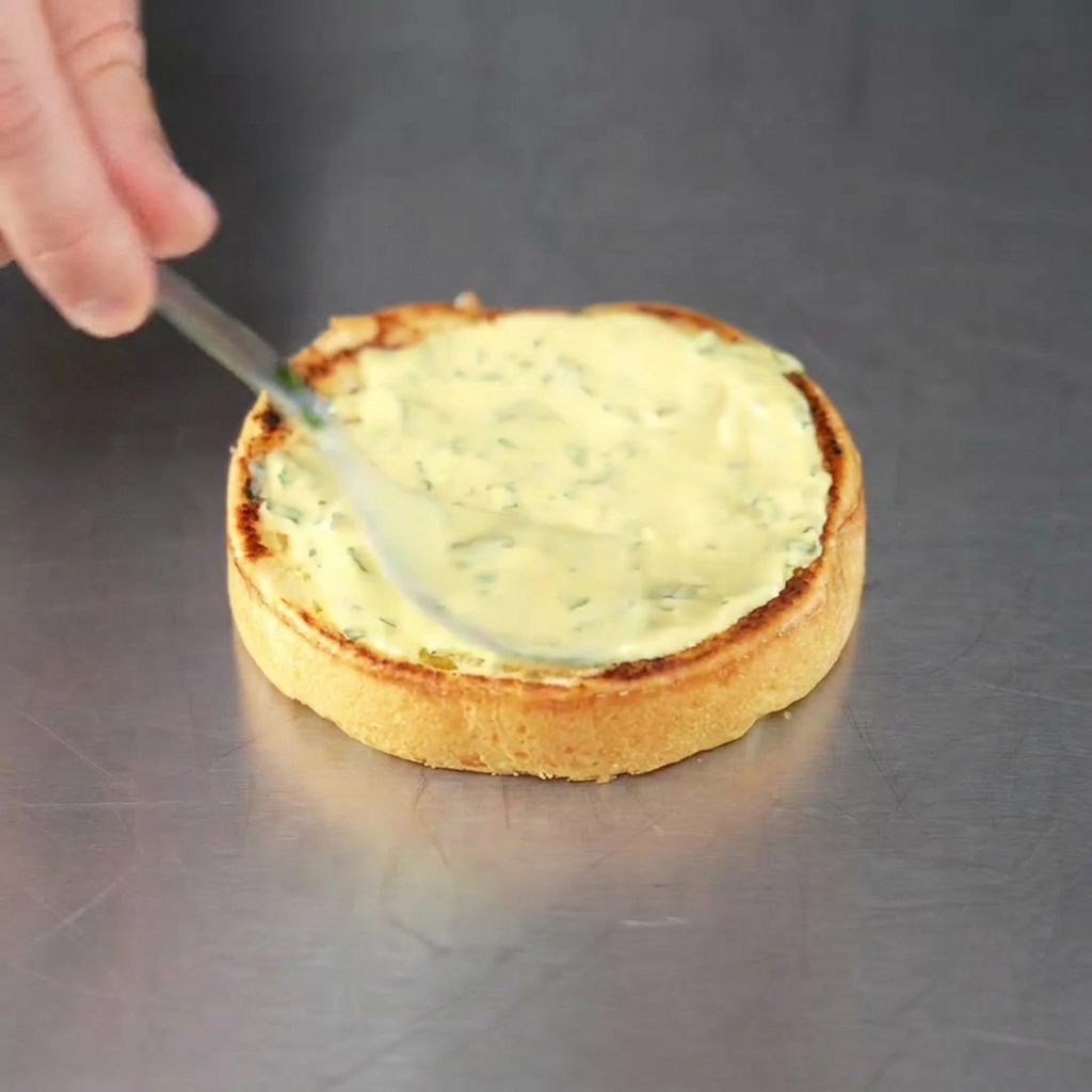 Un panino per hamburger appena tostato su cui viene spalmata la salsa verde alle erbe, pronto per il medaglione di carne.