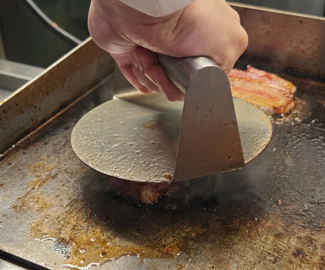 Una pressa per hamburger in metallo che schiaccia una pallina di macinato Wagyu su una superficie calda.