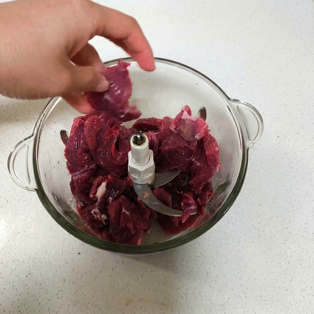 Una mano che mette pezzi di manzo rosso crudo nel contenitore di vetro di un robot da cucina.