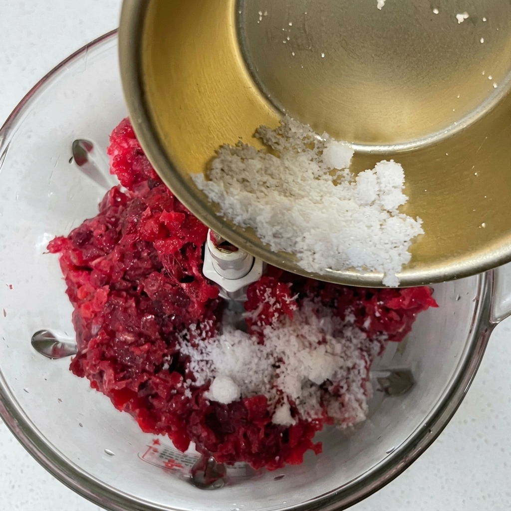 Versare una miscela in polvere bianca di spezie e fecola da una ciotolina di metallo in un robot da cucina con la carne macinata.