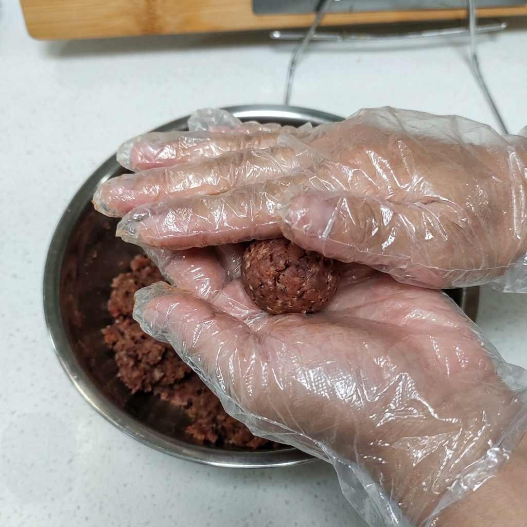 Mani guantate che tengono e premono una pallina di carne macinata condita per formare un burger.