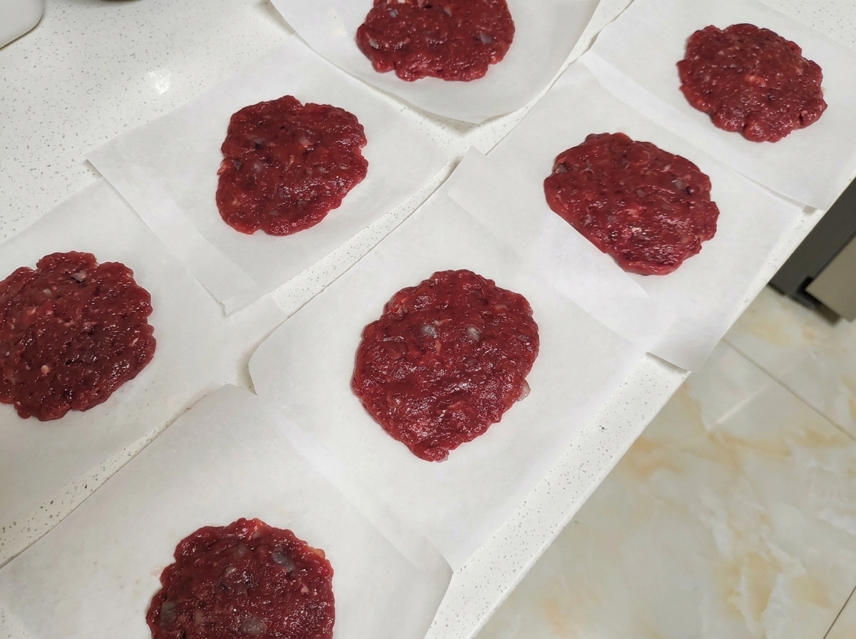 Diversi burger di manzo crudi posizionati singolarmente su quadrati di carta forno su un piano di lavoro chiaro.