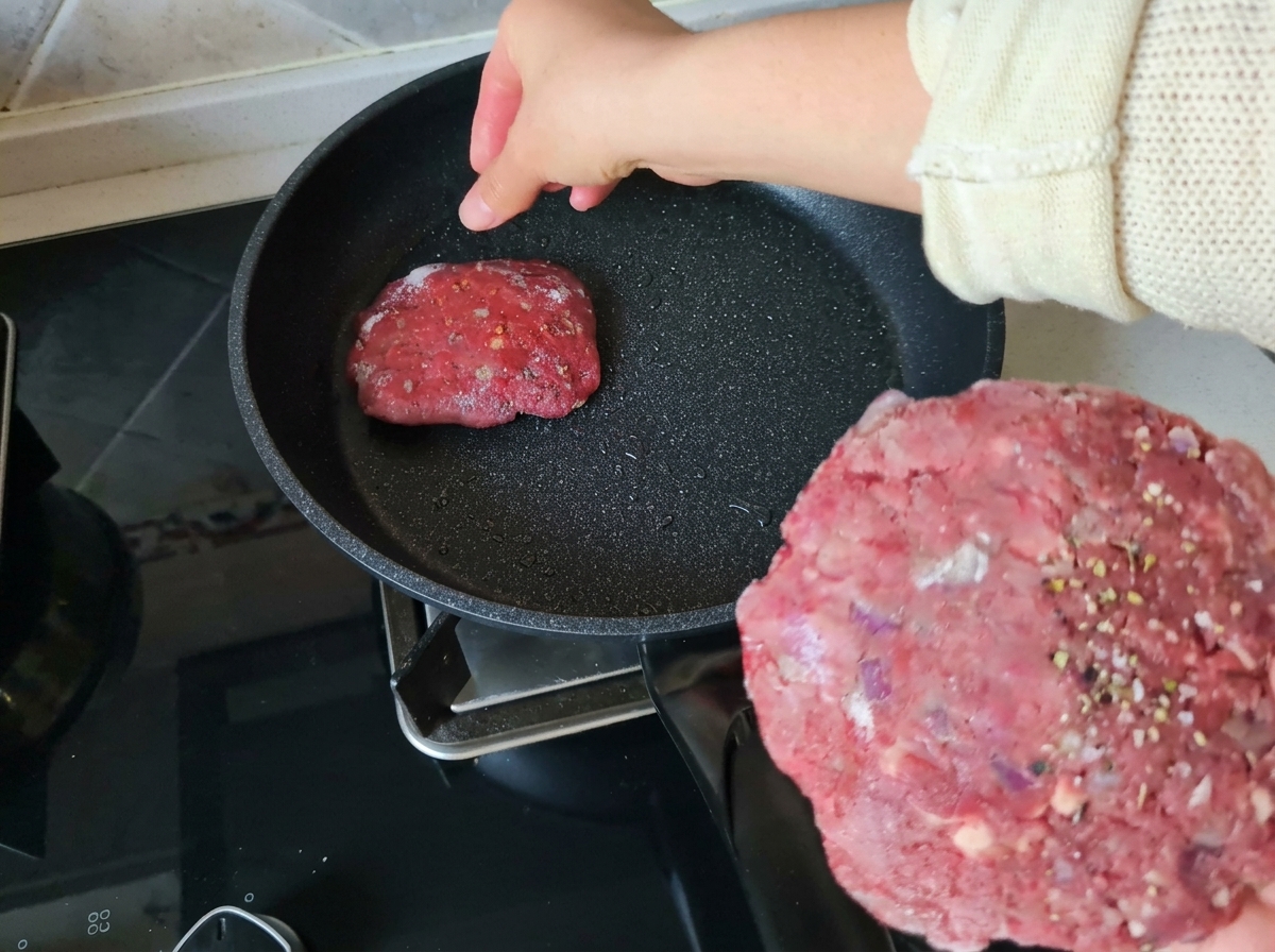 Una mano che tiene un burger di manzo crudo condito mentre ne posiziona un altro in una padella antiaderente leggermente unta.
