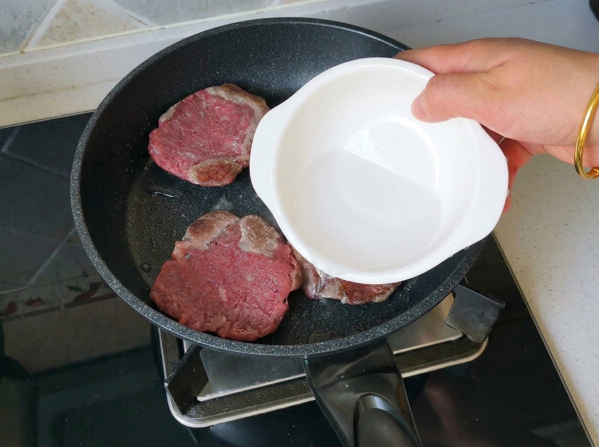 Una mano che versa un goccio dacqua da una ciotolina bianca in una padella contenente tre burger di manzo ben rosolati.
