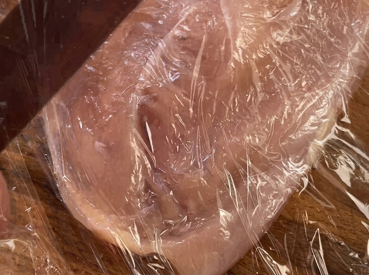 Un mattarello che preme su un petto di pollo coperto con pellicola trasparente.