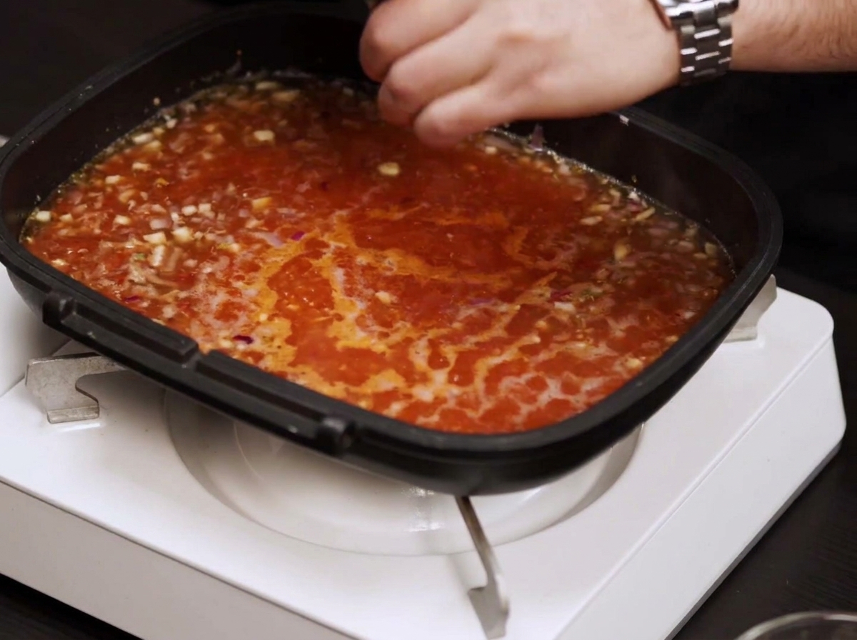 Una padella piena di salsa al pomodoro che sobbolle mentre una mano la insaporisce con sale e pepe.
