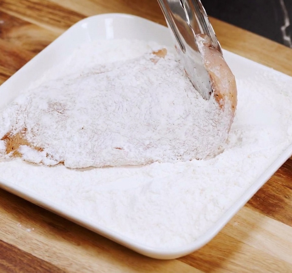 Un petto di pollo crudo infarinato su un piatto rettangolare bianco usando delle pinze da cucina.