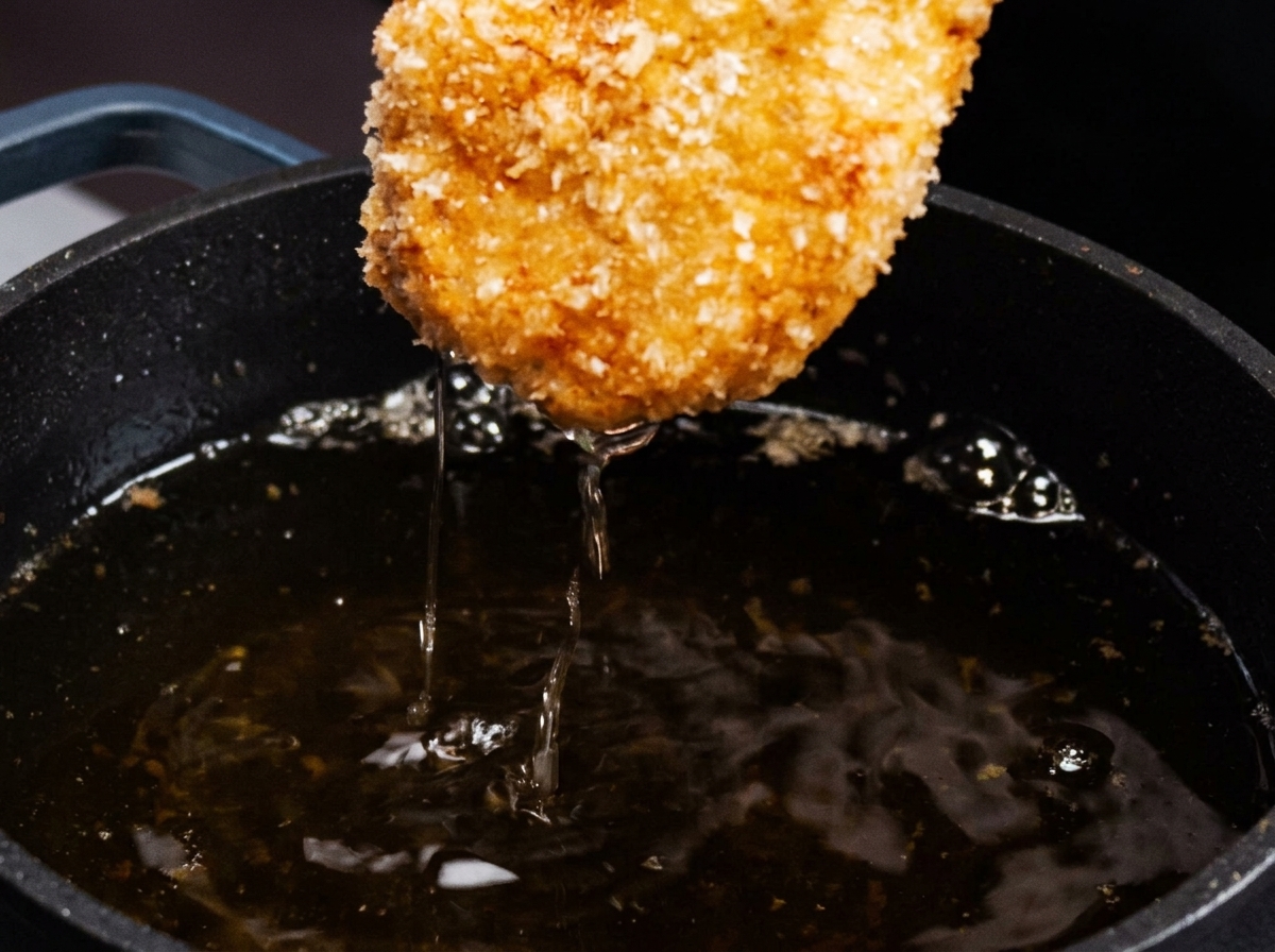Una cotoletta di pollo impanata che viene sollevata da una padella di olio caldo, mostrando una crosticina dorata.