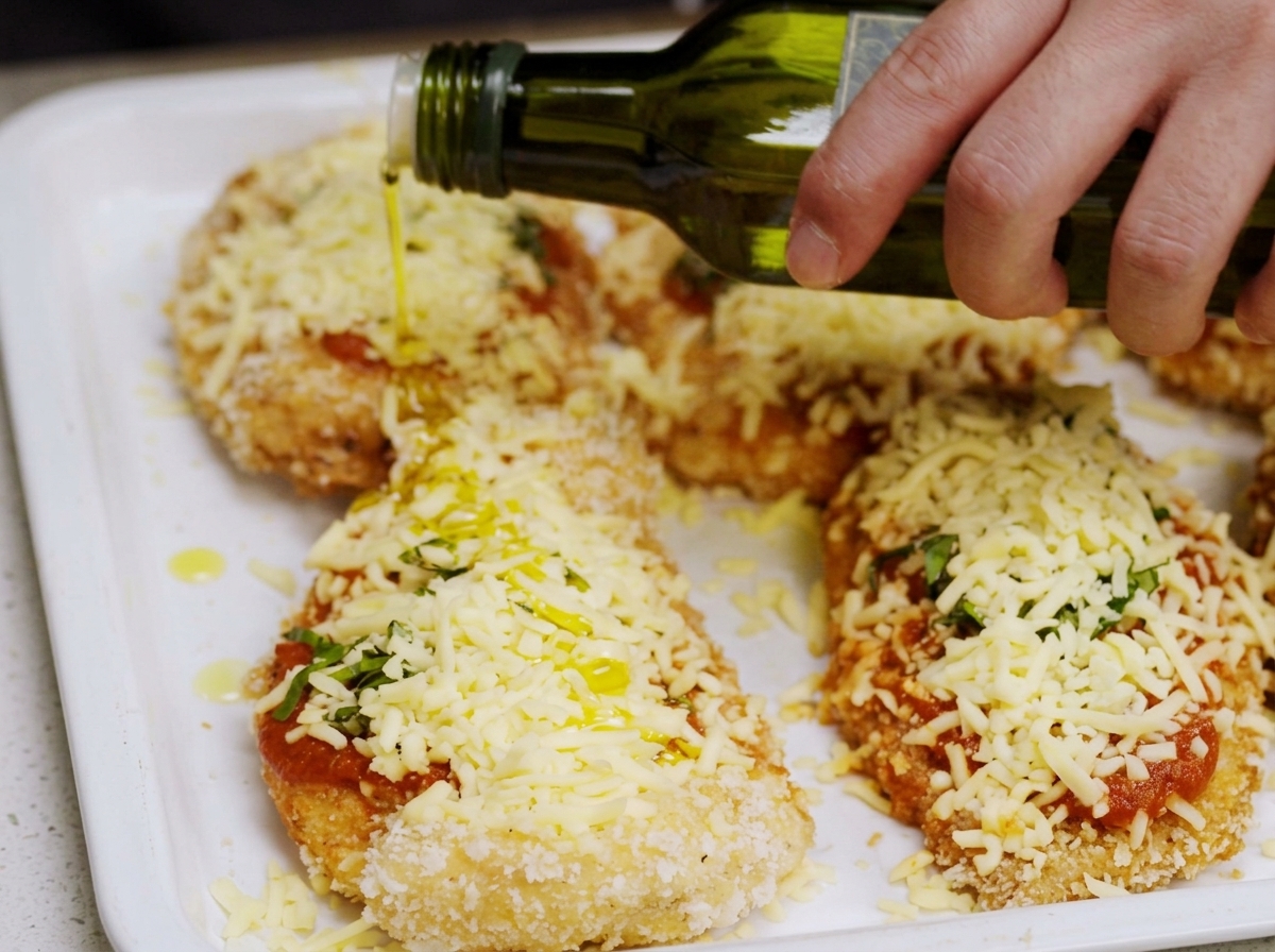 Olio doliva versato da una bottiglia sopra la parmigiana di pollo assemblata sulla teglia.