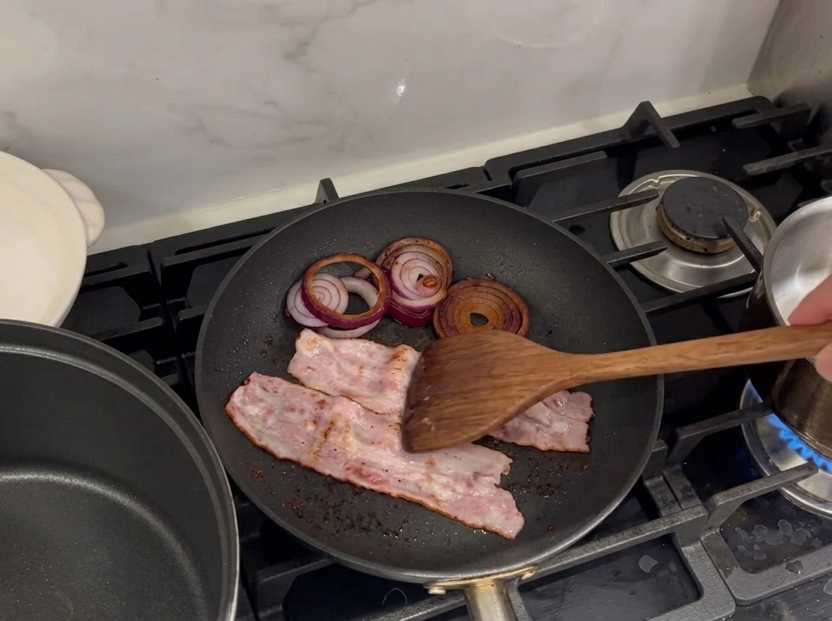 Strisce di pancetta sfrigolanti e anelli di cipolla rossa che friggono in padella.