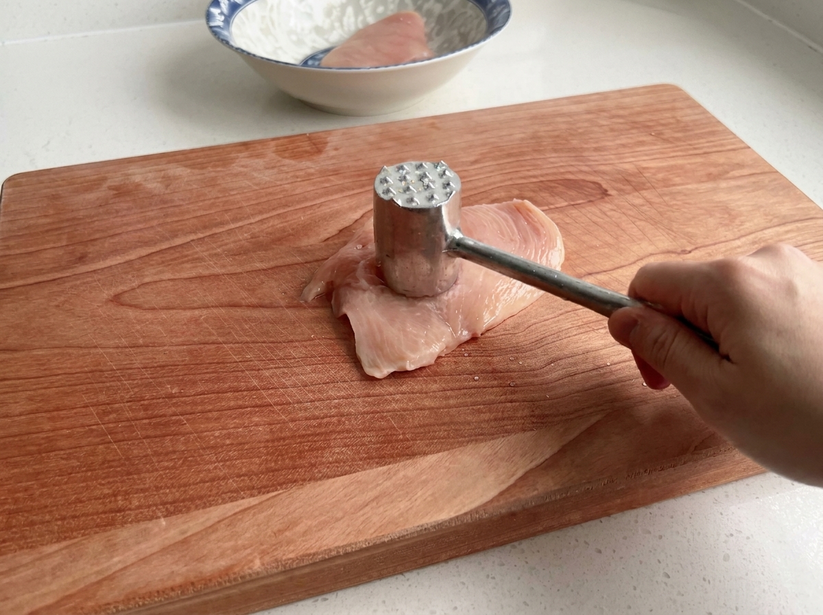 Un primo piano di un batticarne che ammorbidisce un petto di pollo crudo su una superficie di legno.